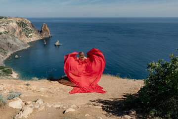 Wall Mural - A woman in a red dress stands on a rocky beach, with the ocean in the background. The scene is serene and peaceful, with the woman's flowing dress adding a sense of grace and beauty to the landscape.