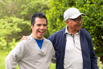 A man and a young man with Down syndrome are standing happily in a park. The young man is wearing a gray sweater.