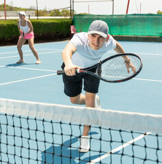 Wall Mural - Portrait of emotional young guy playing tennis on open court in summer, swinging racket to return ball over net. Sportsman ready to hit volley