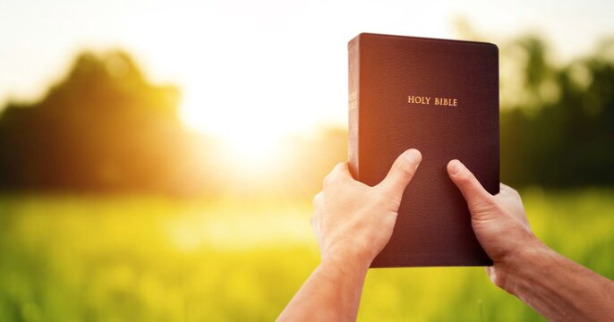 Christian holds bible book in hands with beautiful sunset.
