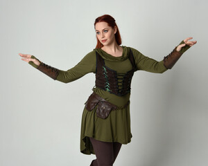 Wall Mural - Close up portrait of beautiful  red haired female model, wearing green and brown medieval fantasy costume with tunic and armour. Standing  with gestural hand poses, isolated on white studio background