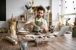 © sofiko14 - Bearded male seated at wooden table with various tools, materials, and laptop in bright well-decorated room. Smiling man working with clay in cozy home pottery studio.