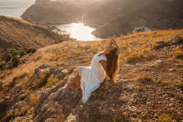 Wall Mural - A woman in a white dress is sitting on a rock overlooking a body of water. She is enjoying the view and taking in the scenery.
