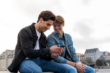 denmark, copenhagen, two young men sitting on a bench using cell phone