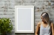 © juliars - Pensive woman looks at an empty white picture frame against a textured brick wall, space for design