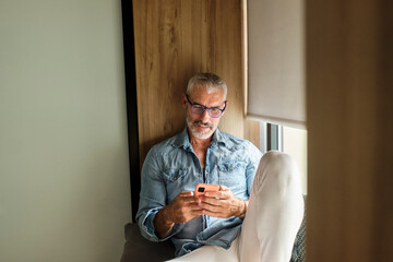 Sticker - Smiling mature man using mobile phone in living room at home