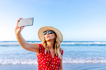 Blond woman wearing red dress and hat and using smartphone and taking a selfie at the beach, making a kissing mouth