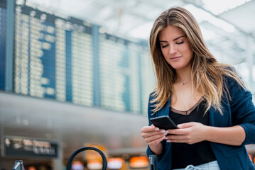 Wall Mural - Young woman using cell phone at departure board looking around