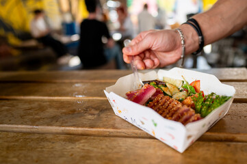 Wall Mural - Close-up of hand holding fork, picking up tuna from a paper bowl of assorted sushi and fresh vegetables, with a blurred restaurant background