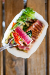 Wall Mural - Close-up of hand holding fork, picking up tuna from a paper bowl of assorted sushi and fresh vegetables, with a blurred restaurant background