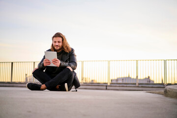 Portrait of bearded young man sitting on parking level at twilight using digital tablet