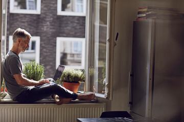 Mature man sitting at the window at home using laptop