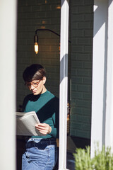 Wall Mural - Woman standing at the window at home reading newspaper