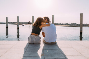 Poster - Rear view of young couple sitting on a pier at the sea kissing