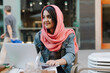© tunedin - Smiling young woman with laptop wearing headscarf at a pavement cafe