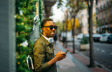 Poster - Young man wearing sunglasses using smart phone while standing on sidewalk in city