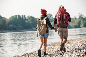 Poster - Rear view of young couple with backpacks walking hand in hand at the riverside