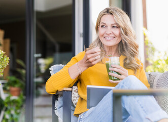 Wall Mural - Portrait of smiling mature woman sitting on terrace with digital tablet enjoying green smoothie