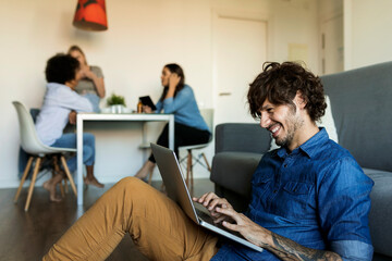 Wall Mural - Smiling man sitting on floor using laptop with friends in background