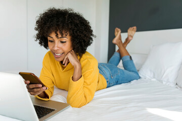 Wall Mural - Smiling woman lying on bed using cell phone and laptop