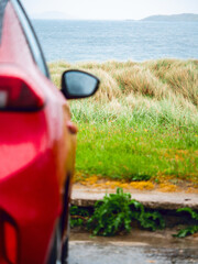 Wall Mural - a red car is parked on the side of a road next to a grassy field with tall grass on a dune and ocean