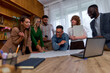 © DusanJelicic - A group of six diverse professionals having a discussion around a table in an office. They are looking at documents and a laptop, appearing engaged and collaborative.