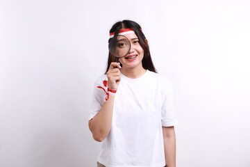 Funny beautiful Asian woman wearing red white flag ribbon, standing while checking something using a magnifying glass, isolated on a white, celebrate Indonesia independence day