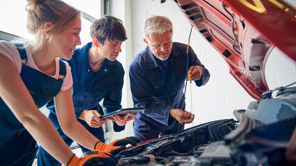 Team Of Car Auto Mechanics With Trainees Looking Under Bonnet Of Car Using Digital Tablet