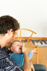 man sitting at home tries to comfort his 6-month-old blond baby who cries inconsolably in his father's arms. Copy space.