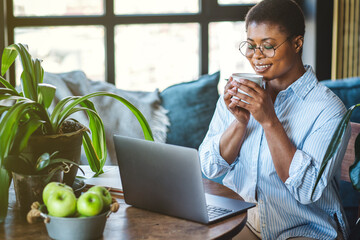 Freelancer morning routine before working day at home. Calm music, coffee, meditation practice. Mindfulness, mental health, calm and happy African American woman at her desk with laptop