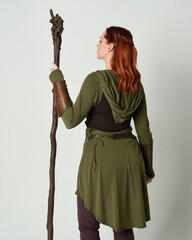 Wall Mural - portrait of red haired female model, wearing green medieval fantasy costume with leather armour. Standing  pose facing away backwards with wood wizard staff weapon, isolated on white studio background