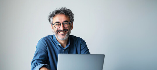 Poster - A man with glasses is sitting at a desk with a laptop in front of him. He is smiling. Man is in his early fifties and has mediterranean features. He is wearing casual outfit. Plain white background