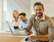 © OceanProd - Startup handsome beard man with team member smiling at camera in office smartphone in hand