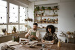 © sofiko14 - Bearded mature man and woman creating pottery pieces in well-lit studio with natural light and green plants. Man sculpts vase while woman rolls clay.