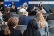 © Anton Gvozdikov - A diverse audience is seen from behind attending a business seminar, listening to speakers on stage discussing various topics.
