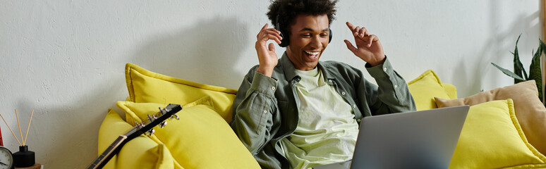 Wall Mural - Young man, African American, sitting on yellow couch, using laptop for online study.