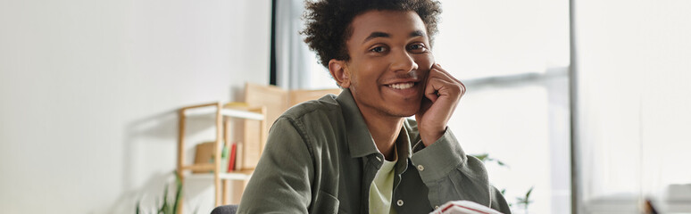 Wall Mural - A young man, African American, sitting at a desk with a book, studying online at home.