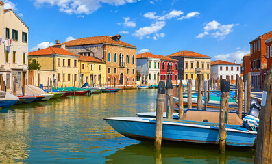Murano Island in Venice, Italy. Boats by the piers and antique italian houses along channel. Blue sky white clouds murano. Sunny summer day. Popular touristic travel destination Europe