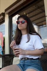 Wall Mural - Young attractive woman in sunglasses sitting in street cafe and holding plastic glass with a juice