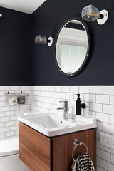 A bathroom sink detail with dark blue walls, subway tiles, a circular mirror, and a floating wood cabinet.