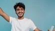 © john - Close up young happy Indian man he wear white t-shirt casual clothes doing selfie shot pov on mobile cell phone do winner gesture isolated on plain pastel light blue cyan background Lifestyle concept
