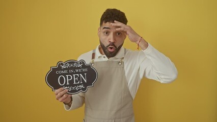 Wall Mural - Young hispanic man in apron, singing openly with amazed face, mouth wide with surprise and disbelief, looking scared over isolated yellow background