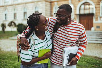 Wall Mural - A young black man and woman share a friendly embrace outside an educational building, both holding study materials, wearing casual attire, and radiating positivity.