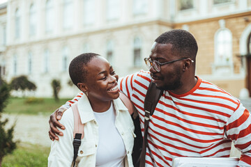 Wall Mural - Two cheerful young black adults share a laugh outside a university building, exemplifying student life with casual attire and books in hand.