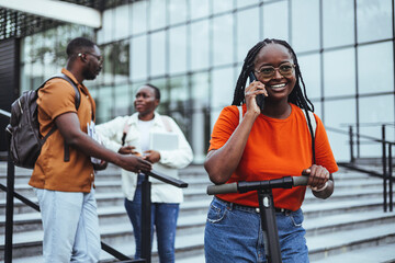 Wall Mural - A joyful black female student engages in a lively phone conversation, leaning on a scooter outside a university building, with fellow students in the background.