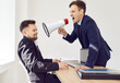 © Studio Romantic - Young angry businessman wearing suit shouting with megaphone on a stressed man company employee working on laptop sitting at the desk on workplace in office. Angry boss yelling at his male worker.