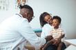 © Dragana Gordic - A diverse male doctor examines a young black boy who is sitting on his mother's lap, both attentive and calm, in a medical office setting with educational posters in the background.