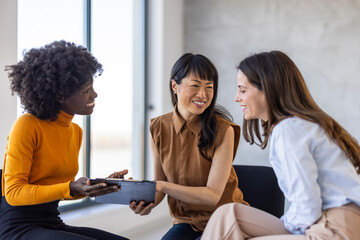 Wall Mural - Three women of varied ethnic backgrounds in professional attire discuss work, sharing insights over a clipboard in a modern office setting.