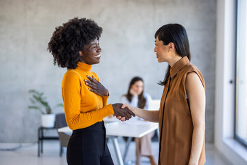 Wall Mural - A cheerful Black businesswoman in a vibrant orange top clasps hands with an Asian colleague wearing a sleeveless brown blouse, exemplifying professional collaboration within a sleek