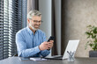 © Liubomir - Smiling mature businessman using smartphone while sitting at office desk with laptop and notebook. He looks happy and productive.
